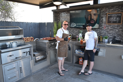 Two men stand in an outdoor kitchen by a Kokomo Grills 32-Inch Built-In Charcoal Grill. One, in apron and gloves, holds tongs; the other has a drink. Behind them are a TV, snacks, and a chalkboard titled “Butcher’s Guide.”.