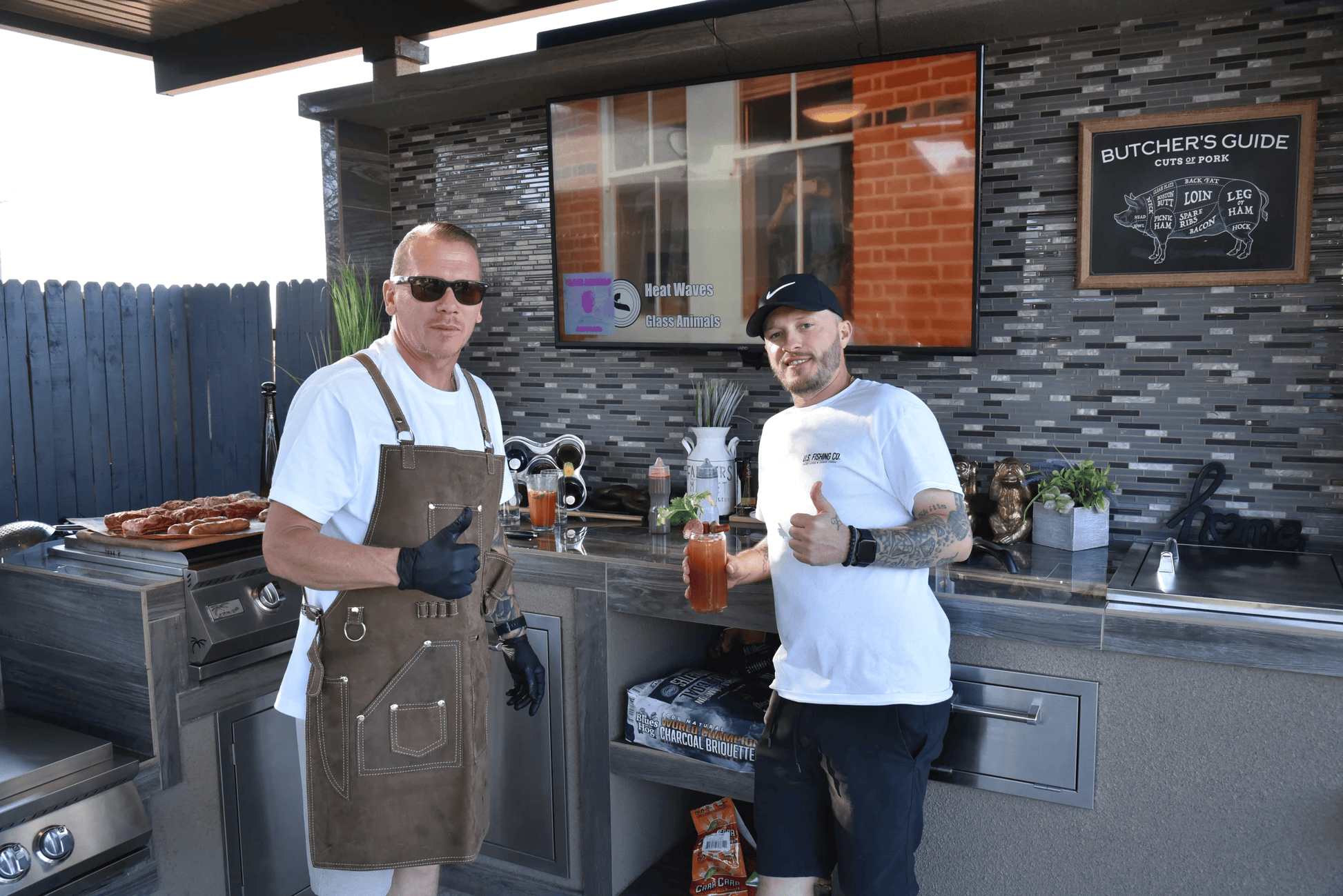 Two men give thumbs up by the Kokomo Grills 32-Inch Built-In Charcoal Grill, with meats cooking, snacks nearby, and a TV plus Butchers Guide sign in the outdoor kitchen. One wears an apron and sunglasses; the other has a cap and holds a drink.