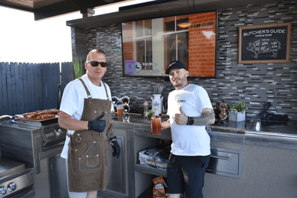 Two men give thumbs up by the Kokomo Grills 32-Inch Built-In Charcoal Grill, with meats cooking, snacks nearby, and a TV plus Butchers Guide sign in the outdoor kitchen. One wears an apron and sunglasses; the other has a cap and holds a drink.