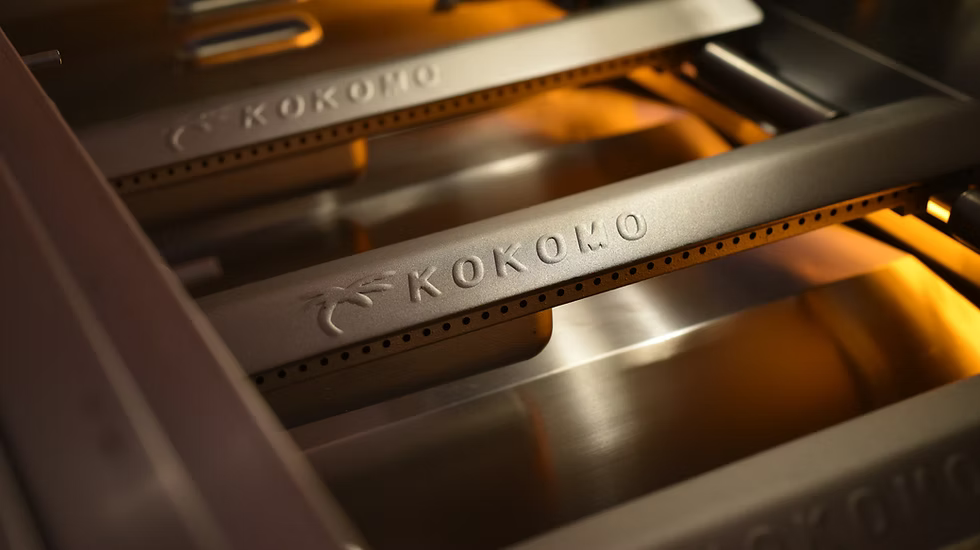 Close-up of Kokomo Grills 32-Inch Elite Built-In Gas Grill (4 Burner) burners, featuring the Kokomo Grills name and palm tree logo engraved, with orange flames glowing—ideal for any outdoor kitchen.