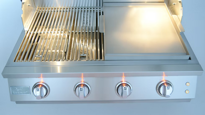 A close-up of the Kokomo Grills 32-Inch Elite Built-In Gas Grill (4 Burner) shows four lit knobs, grill grates on the left, and a flat griddle surface on the right in an outdoor kitchen, with its lid partially open.