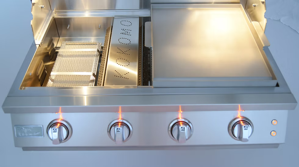 The Kokomo Grills 32-Inch Elite Built-In Gas Grill (4 Burner) by Kokomo Grills includes four lit control knobs, a flat griddle on the right, and grilling and smoking baskets on the left—ideal for outdoor kitchens.