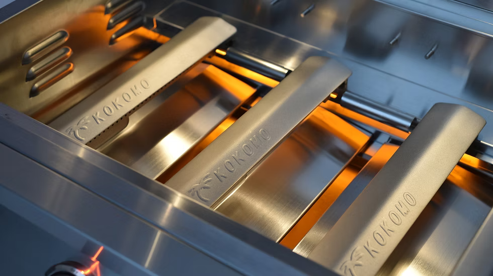 Close-up of the Kokomo Grills 32-Inch Elite Built-In Gas Grill (4 Burner) interior, featuring three KOKOMO-labeled metal heat deflectors and glowing orange flames—a stunning, functional highlight for your outdoor kitchen.