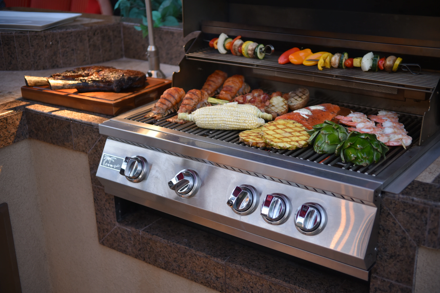 The Kokomo Grills 32-Inch Professional Built-In Gas Grill (4 Burner) by Kokomo Grills is loaded with corn, skewered veggies, shrimp, sausage, artichokes, and fish. A juicy steak sits on a cutting board next to this stunning outdoor kitchen centerpiece.