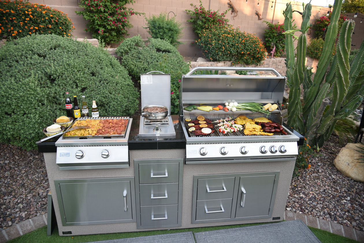 The Kokomo Grills 40-Inch Built-In Gas Grill (5 Burner / Back Burner) by Kokomo Grills anchors a spacious outdoor kitchen, sizzling meats and veggies, with sauces and drinks nearby, all set in a landscaped backyard with flowers and a large cactus.