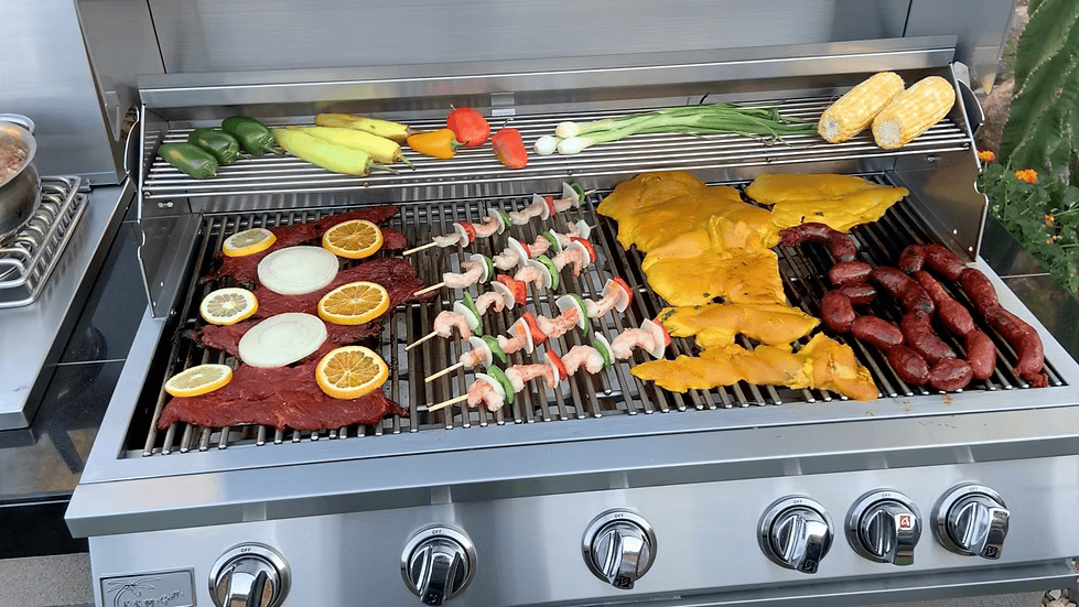 An outdoor kitchen scene features the Kokomo Grills 40-Inch Built-In Gas Grill (5 Burner / Back Burner) from Kokomo Grills, cooking marinated steak with orange and onion, shrimp skewers, chicken, sausages, corn, green onions, peppers, and squash.