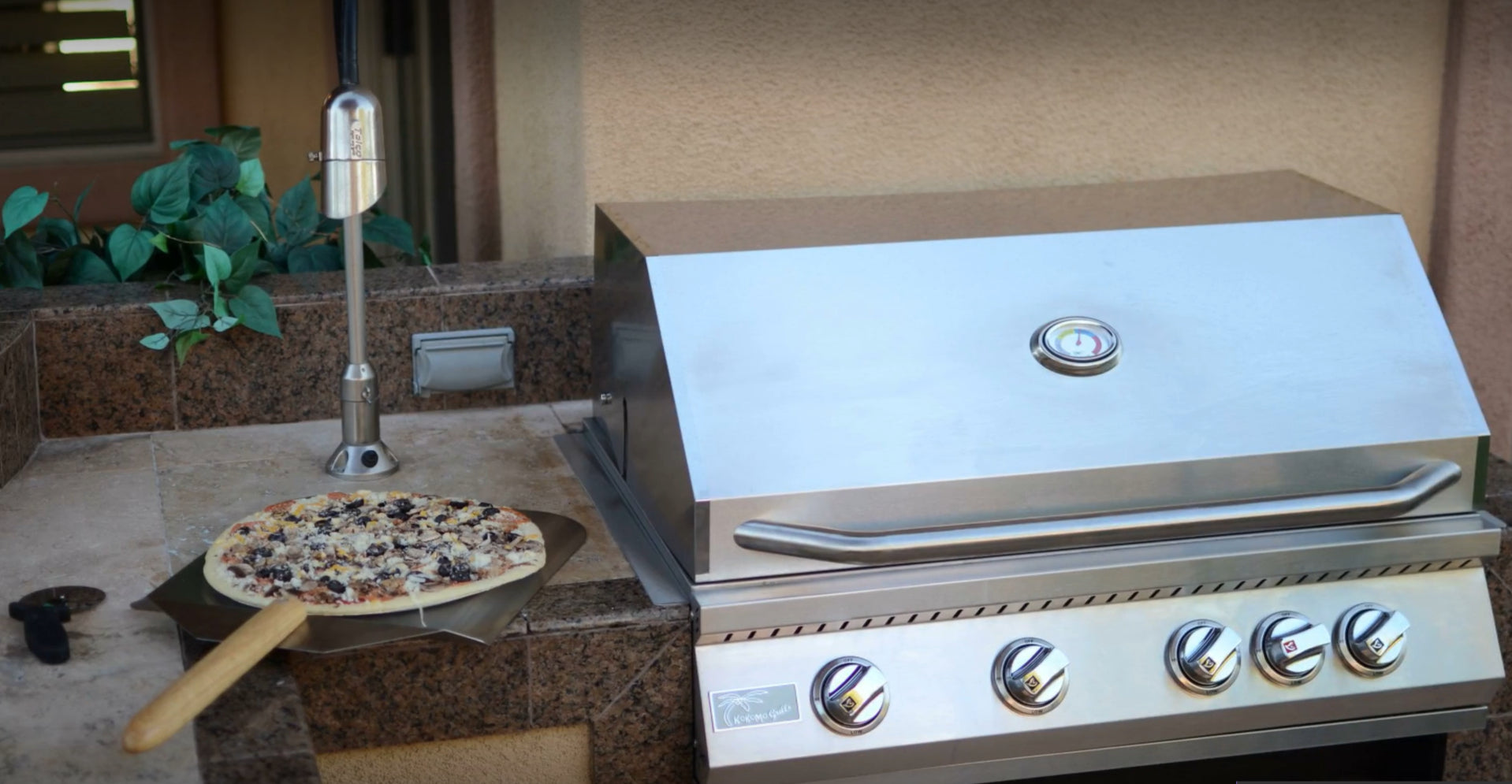 A fresh pizza on a wooden peel is ready to be baked beside the Kokomo Grills 40-Inch Professional Built-In Gas Grill (5 Burner) in an outdoor stone patio kitchen setup.