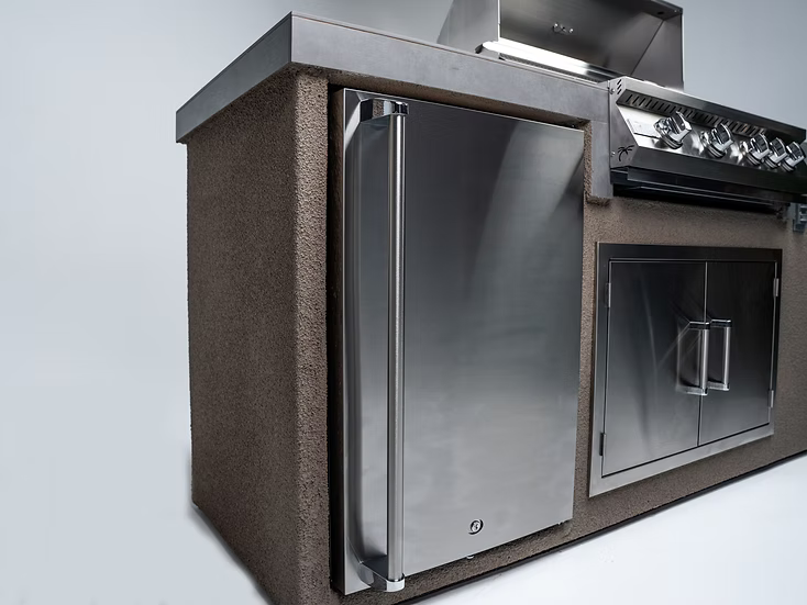 The Kokomo Grills 7-Foot Straight Kitchen Island features a stainless steel fridge, built-in Kokomo Grills with control knobs, and storage cabinets in a textured, stone-like enclosure.