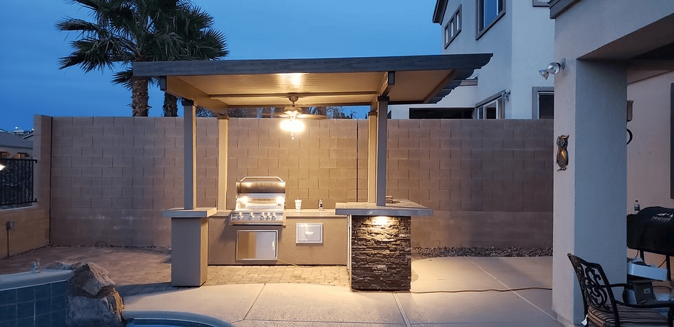An outdoor kitchen features the Kokomo Grills Aruba BBQ Island with a built-in grill beneath a 12'x12' pergola, ceiling light, and is set poolside beside a tall stone and brick privacy wall with palm trees in the background.