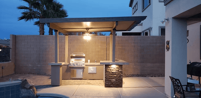 An outdoor kitchen features the Kokomo Grills Aruba BBQ Island with a built-in grill beneath a 12'x12' pergola, ceiling light, and is set poolside beside a tall stone and brick privacy wall with palm trees in the background.