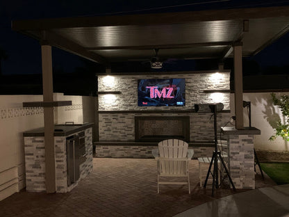 The Kokomo Grills Aruba BBQ Island with 12’x12’ pergola features a covered seating area, stone fireplace, flat-screen TV, armchair facing the TV, modern lighting, and a built-in BBQ grill for stylish outdoor dining and entertaining.
