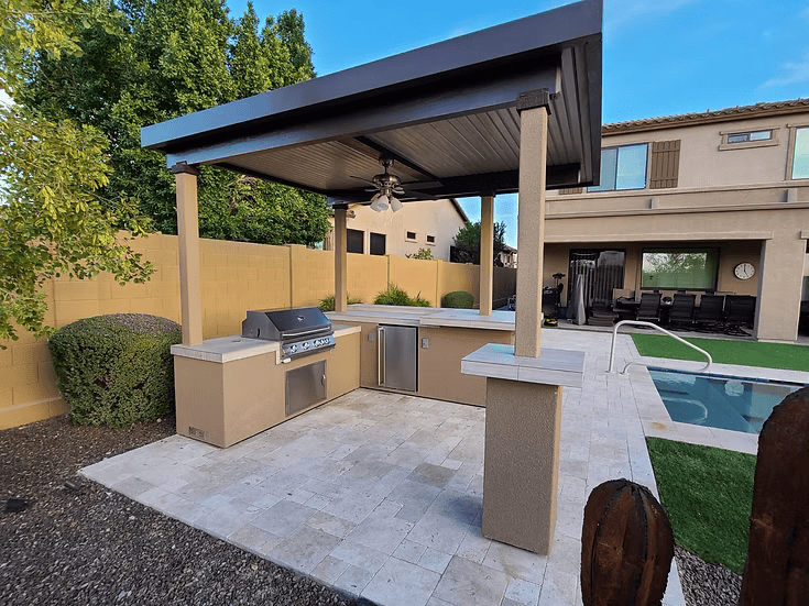 The Kokomo Grills Aruba BBQ Island with 12' x 12' pergola and built-in grill by Kokomo Grills sits on a tiled patio beside the pool, offering modern outdoor cooking with views of trees and a two-story house beyond.