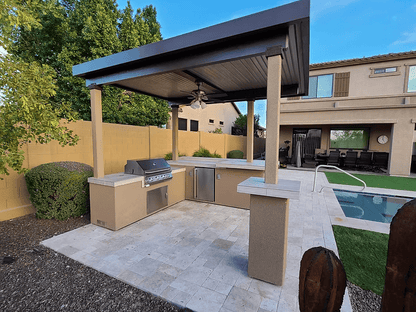 The Kokomo Grills Aruba BBQ Island with 12' x 12' pergola and built-in grill by Kokomo Grills sits on a tiled patio beside the pool, offering modern outdoor cooking with views of trees and a two-story house beyond.