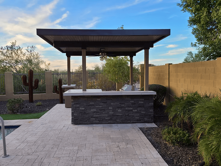 A Kokomo Grills Aruba BBQ Island with built-in BBQ grill sits under a 12x12 ft pergola beside a pool, surrounded by desert cacti, a tall brick privacy wall, and lush green plants.