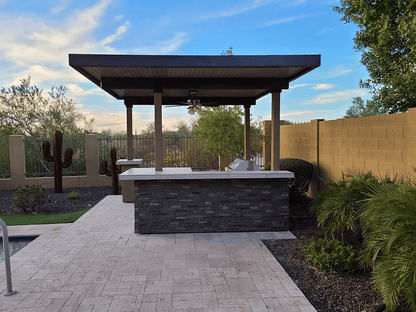 A Kokomo Grills Aruba BBQ Island with built-in BBQ grill sits under a 12x12 ft pergola beside a pool, surrounded by desert cacti, a tall brick privacy wall, and lush green plants.