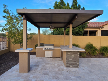 Modern outdoor kitchen with the Kokomo Grills Aruba BBQ Island, set under a 12x12 ft pergola with built-in BBQ grill by Kokomo Grills, stone countertops, planter, tiled patio, and landscaping along a yellow brick wall.