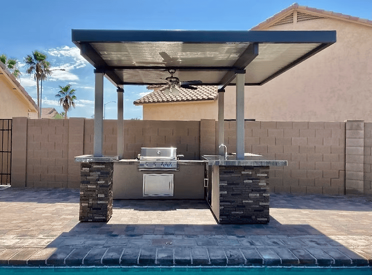 The Kokomo Grills Aruba BBQ Island features a built-in grill, stone countertops, sink, and ceiling fan under a 12x12-foot metal pergola. It sits poolside and is enclosed by a tan brick privacy wall.