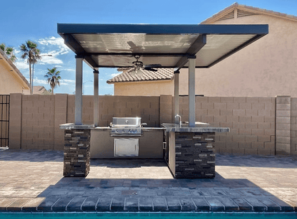 The Kokomo Grills Aruba BBQ Island features a built-in grill, stone countertops, sink, and ceiling fan under a 12x12-foot metal pergola. It sits poolside and is enclosed by a tan brick privacy wall.