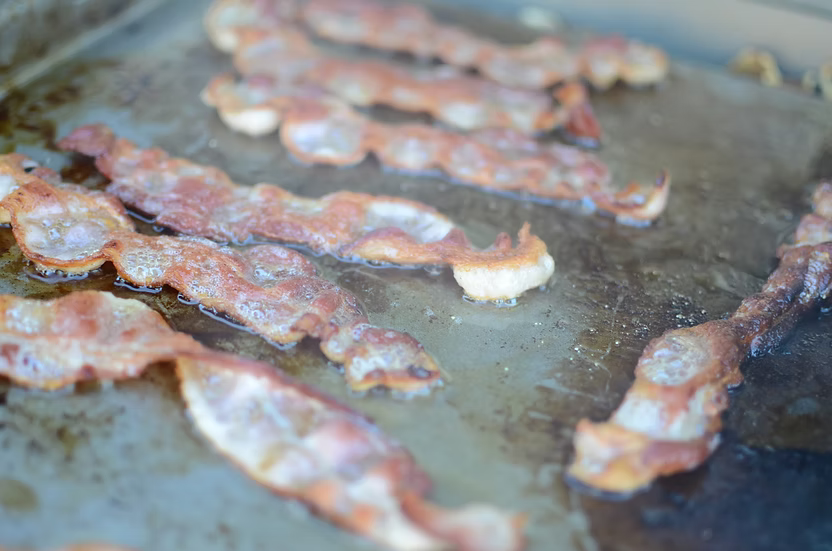 Crispy bacon strips sizzling on the Kokomo Grills BBQ Grill Griddle Plate Insert, with oil and browned spots visible on the stainless steel surface.