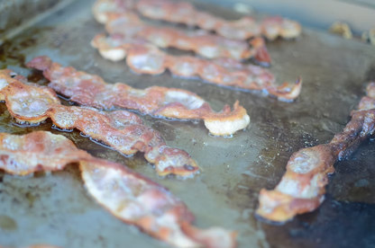 Crispy bacon strips sizzling on the Kokomo Grills BBQ Grill Griddle Plate Insert, with oil and browned spots visible on the stainless steel surface.