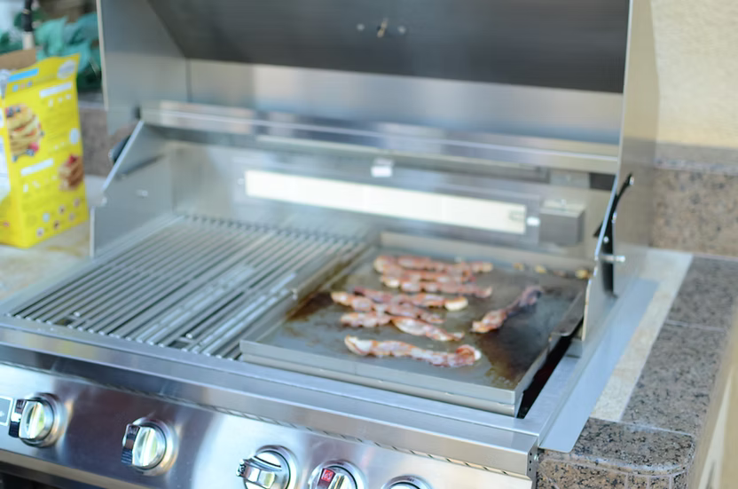 Bacon sizzles on a Kokomo Grills BBQ Grill Griddle Plate Insert, a stainless steel accessory for outdoor grills. The grill lid is open with steam rising, and a yellow food box appears in the background on the left.