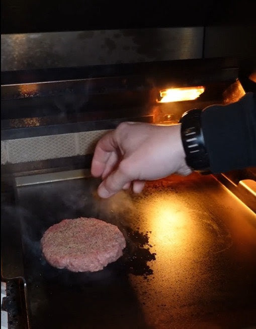 A hand sprinkles seasoning onto a raw burger patty sizzling on the Kokomo Grills BBQ Grill Griddle Plate Insert by Kokomo Grills, with backlighting illuminating the scene.
