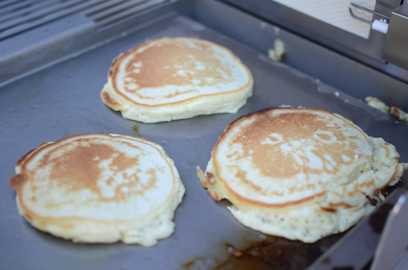 Three golden brown pancakes cook on a Kokomo Grills BBQ Grill Griddle Plate Insert, their edges slightly crispy with some spilled batter nearby. Each pancake is at a different stage of cooking, making it ideal for a perfect breakfast.