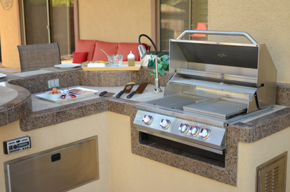 A modern outdoor kitchen features a Kokomo Grills BBQ Grill Griddle Plate Insert in a stainless steel grill, with utensils and food prep items on a granite countertop. A red cushioned seat and windowed wall are visible in the background.