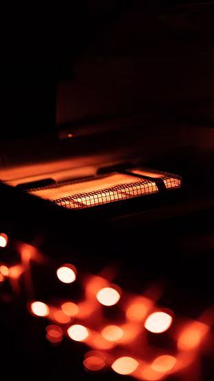 A close-up of the Kokomo Grills BBQ Grill Infrared Sear Zone Burner KO-IRSB shows glowing heating coils casting a warm orange light from the stainless steel Kokomo Grills unit, with blurred orange highlights in a dimly lit room.