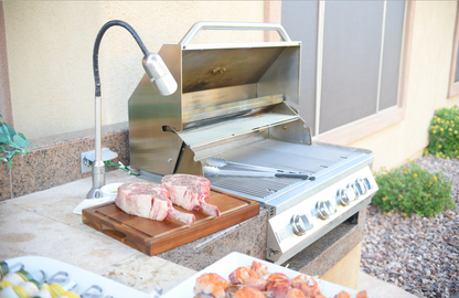 A Kokomo Grills BBQ Grill Infrared Sear Zone Burner KO-IRSB sits open on a patio beside two raw tomahawk steaks on a wooden board, plus skewers and grilled food on a tray. Grilling tongs rest on the brand’s signature burner.