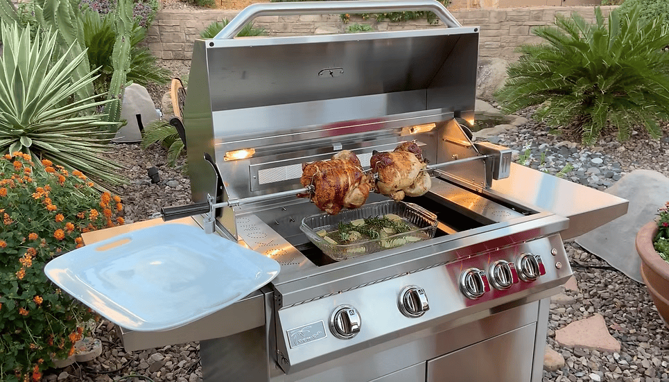 A Kokomo Grills BBQ Grill Rotisserie Kit by Kokomo Grills slowly roasts two chickens and a tray of vegetables on a stainless steel outdoor grill, set in a backyard with lush plants, stones, and a brick wall in the background.