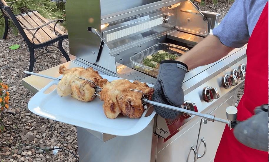 Wearing a red apron and oven mitts, a person holds a tray with two cooked chickens beside an outdoor grill using the Kokomo Grills BBQ Grill Rotisserie Kit. Grilled vegetables are seen inside the open BBQ grill, with a bench and plants in the background.