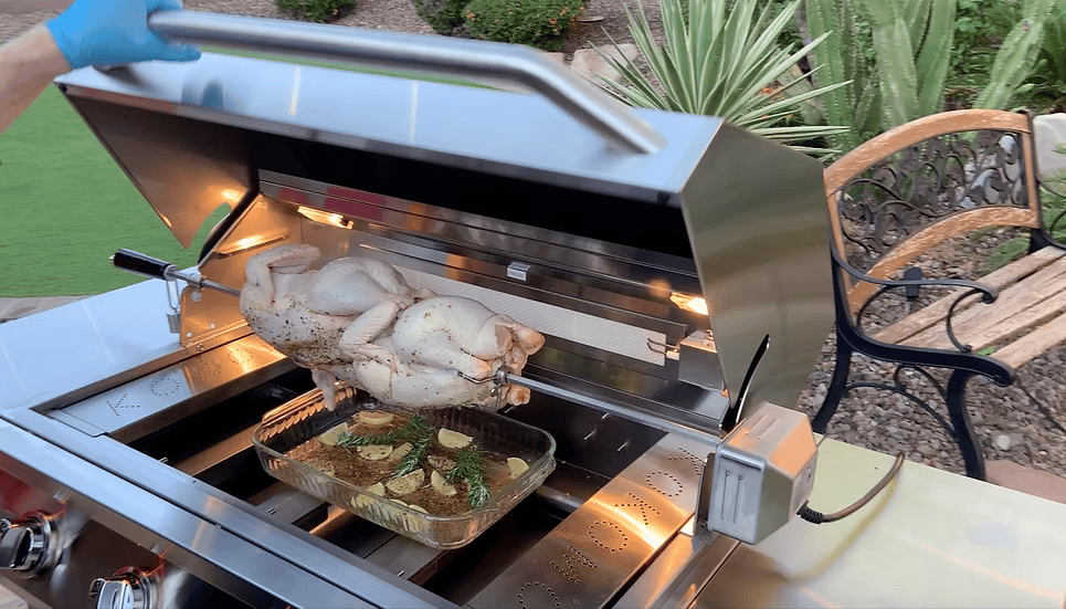 A whole chicken roasts on a Kokomo Grills BBQ Grill Rotisserie Kit inside an outdoor grill, while vegetables cook below. A person in a blue glove lifts the lid, with patio furniture and plants in the background.