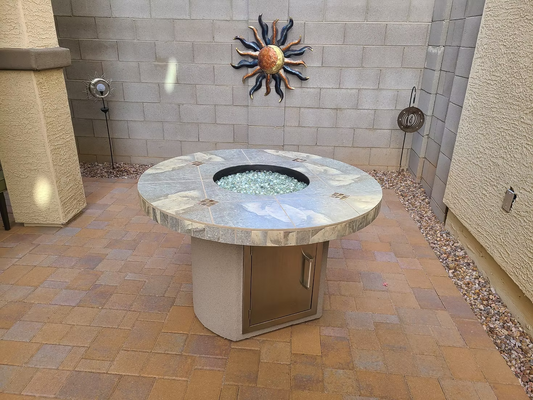 A Kokomo Grills Bahama Outdoor Fire Table by Kokomo Grills, featuring glass rocks, is set on a paved patio beside a stone wall adorned with a metal sun and garden ornaments, with small stones lining the wall’s edge.