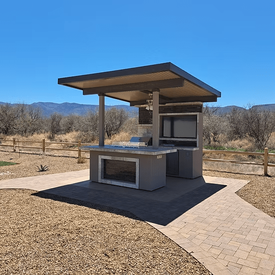 The Kokomo Grills Baja Kitchen Island with Patio Cover Top by Kokomo Grills sits on a paved area bordered by mulch, with a wooden fence, dry grass, leafless trees, and mountains in the background under a clear blue sky.