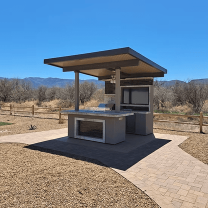 The Kokomo Grills Baja Kitchen Island with Patio Cover Top by Kokomo Grills sits on a paved area bordered by mulch, with a wooden fence, dry grass, leafless trees, and mountains in the background under a clear blue sky.