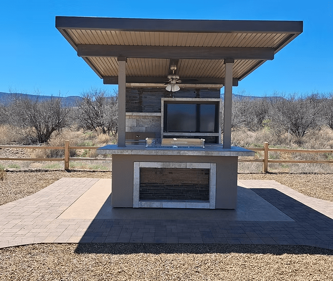 The Kokomo Grills Baja Kitchen Island with Patio Cover Top by Kokomo Grills features a TV, ceiling fan, and built-in grill BBQ island on paved ground with a wooden fence and dry grass landscape under a clear blue sky.