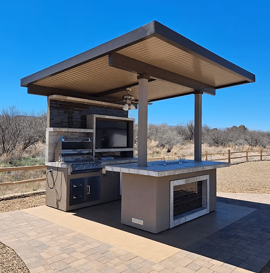 The Kokomo Grills Baja Kitchen Island with Patio Cover Top by Kokomo Grills features a BBQ island with grill, sink, and countertop, plus a TV, all set on a paved patio surrounded by nature under a clear blue sky.