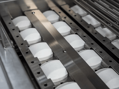 Rows of white, oval-shaped tablets moving along a stainless steel conveyor evoke the precision seen in assembling the Kokomo Grills Baja with Fire 7-Foot 6-Inch by 9-Foot Kitchen Island, where top-quality materials from Kokomo Grills are essential.