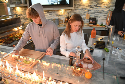 A young man and woman prepare food and drinks at a Kokomo Grills Best Outdoor Kitchen Bermuda BBQ Island with TV, working at the counter near a flame feature surrounded by grilled food, an orange, a decanter, and bartending tools.