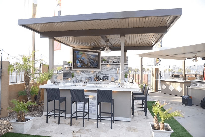 The Kokomo Grills Best Outdoor Kitchen Bermuda BBQ Island with TV by Kokomo Grills features a tile countertop, bar stools, and sits under a modern pergola with palm plants, tiled flooring, a gate, and extra patio space surrounding the area.