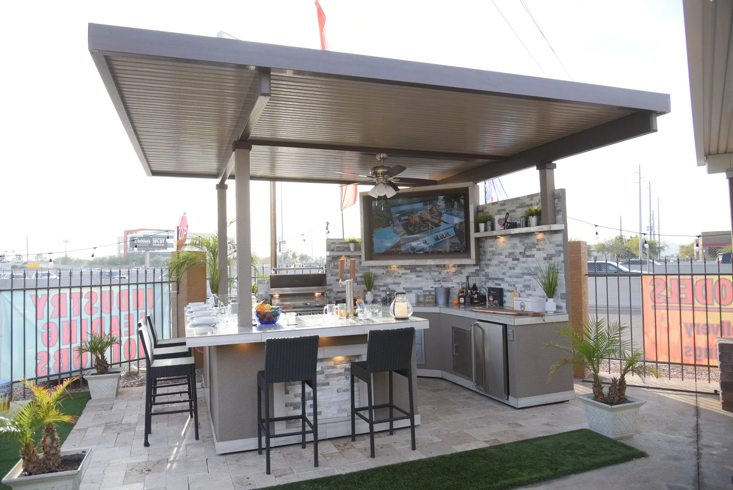 Chic outdoor kitchen under a metal pergola showcasing the Kokomo Grills Best Outdoor Kitchen Bermuda BBQ Island with TV, bar seating, grill, sink, modern decor, and barstools—plus potted plants and views of signs and buildings beyond the fenced area.