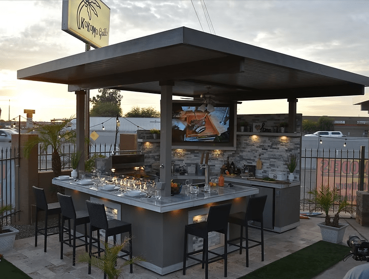 The Kokomo Grills Best Outdoor Kitchen Bermuda BBQ Island with TV features a roof, bar seating, countertop appliances, and sits on stone tile with palm plants and a metal fence; a street is visible in the background.
