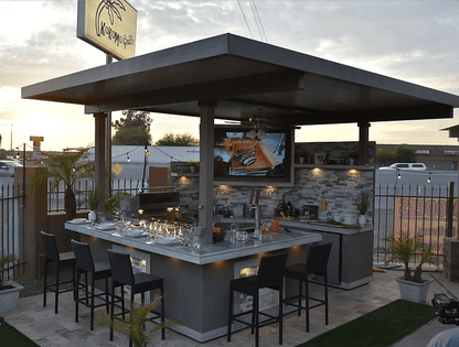 The Kokomo Grills Best Outdoor Kitchen Bermuda BBQ Island with TV features a roof, bar seating, countertop appliances, and sits on stone tile with palm plants and a metal fence; a street is visible in the background.