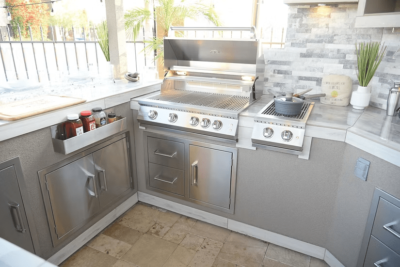 A stylish BBQ island setup featuring the Kokomo Grills Best Outdoor Kitchen Bermuda BBQ Island with TV, stainless steel grill, side burner with pot, drawers, white countertops, potted plants, and a tiled backsplash.