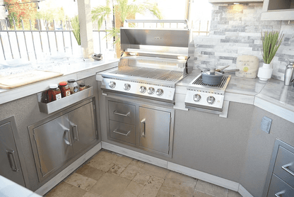 A stylish BBQ island setup featuring the Kokomo Grills Best Outdoor Kitchen Bermuda BBQ Island with TV, stainless steel grill, side burner with pot, drawers, white countertops, potted plants, and a tiled backsplash.