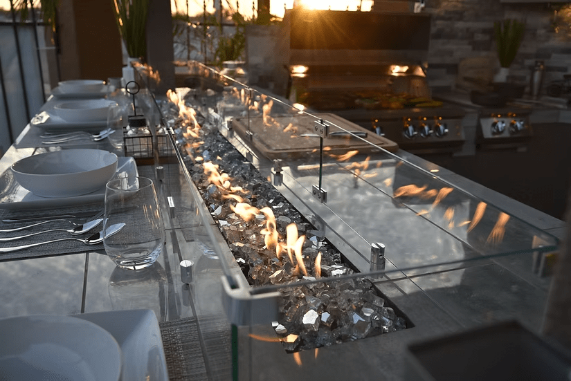A modern outdoor dining table with plates and glasses sits beside a glass fire pit. In the background, a Kokomo Grills Best Outdoor Kitchen Bermuda BBQ Island with TV from Kokomo Grills sizzles with food at sunset.