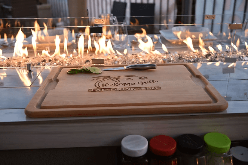 A wooden cutting board with lime slices and a knife sits in front of the Kokomo Grills Best Outdoor Kitchen Bermuda BBQ Island with TV, perfect for any outdoor space. The board displays the Kokomo Grills logo, with condiment jars below.