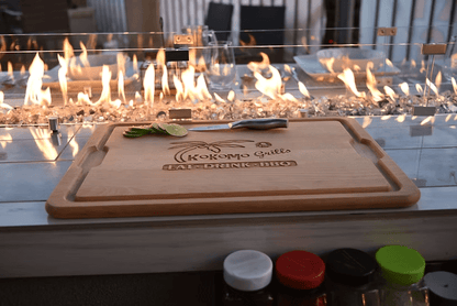 A wooden cutting board with lime slices and a knife sits in front of the Kokomo Grills Best Outdoor Kitchen Bermuda BBQ Island with TV, perfect for any outdoor space. The board displays the Kokomo Grills logo, with condiment jars below.