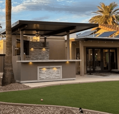 The Kokomo Grills Best Outdoor Kitchen Bermuda BBQ Island with TV sits beneath a lit flat roof, near a home with large windows and solar panels, surrounded by palm trees and artificial grass at sunset.